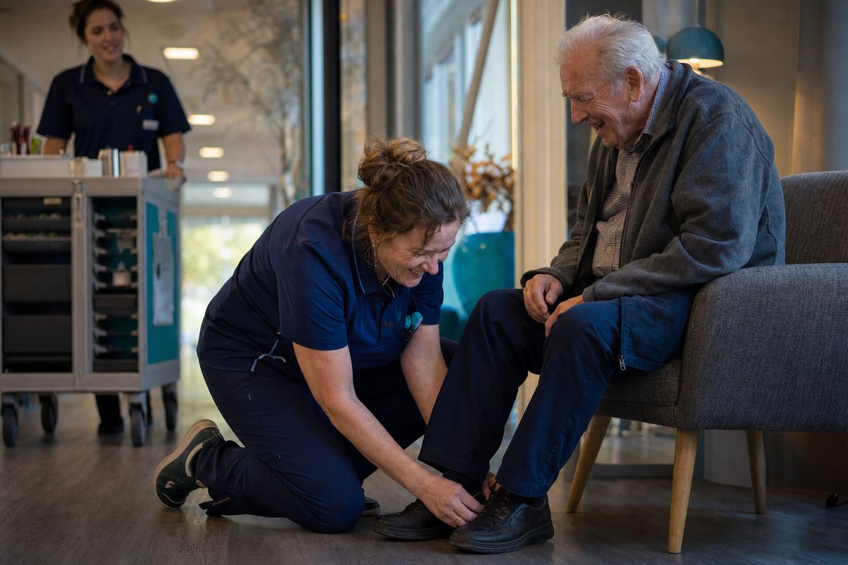 Een zorgmedewerker helpt een oudere bewoner met zijn schoenen